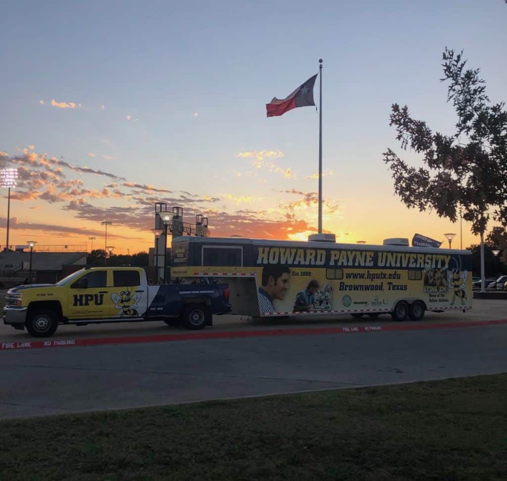 A Howard Payne University bus parked near a field under a sunset sky with the Howard Payne University flag flying in the background. | HPU