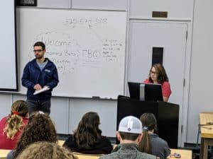 Two people present at the front of a Howard Payne University classroom with students looking on, featuring a whiteboard with meeting information in the background. | HPU