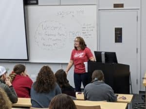 A woman presenting at a Howard Payne University gamma beta phi meeting in front of an audience with welcome information written on a whiteboard. | HPU