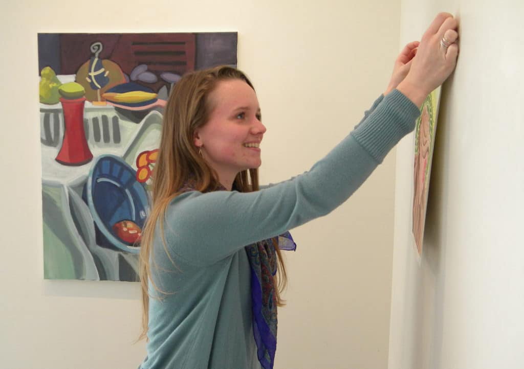 Woman hanging a picture on a white wall with colorful artwork from Howard Payne University in the background. | HPU