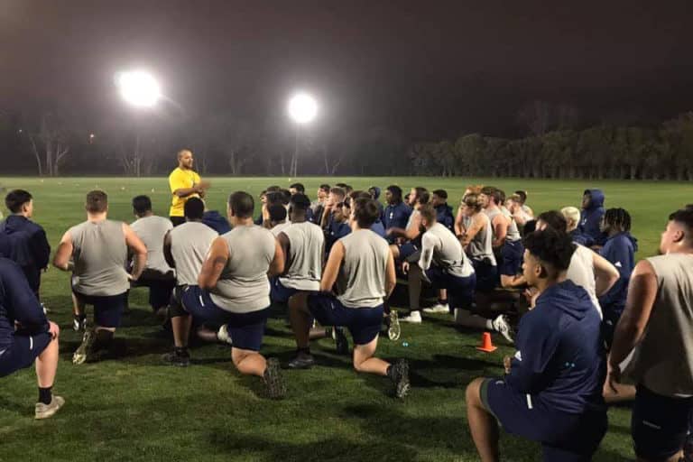A coach addressing a group of Howard Payne University athletes during a nighttime practice session on the field. | HPU