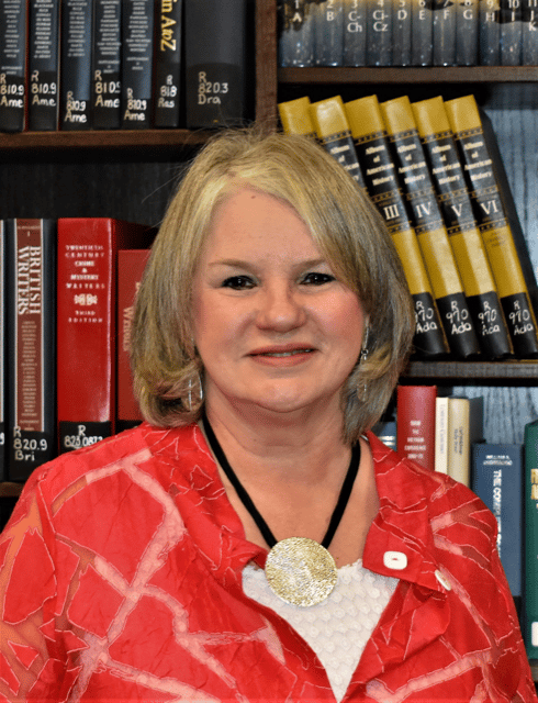 Woman in a red blouse with a Howard Payne University medallion necklace, smiling in front of a bookshelf. | HPU
