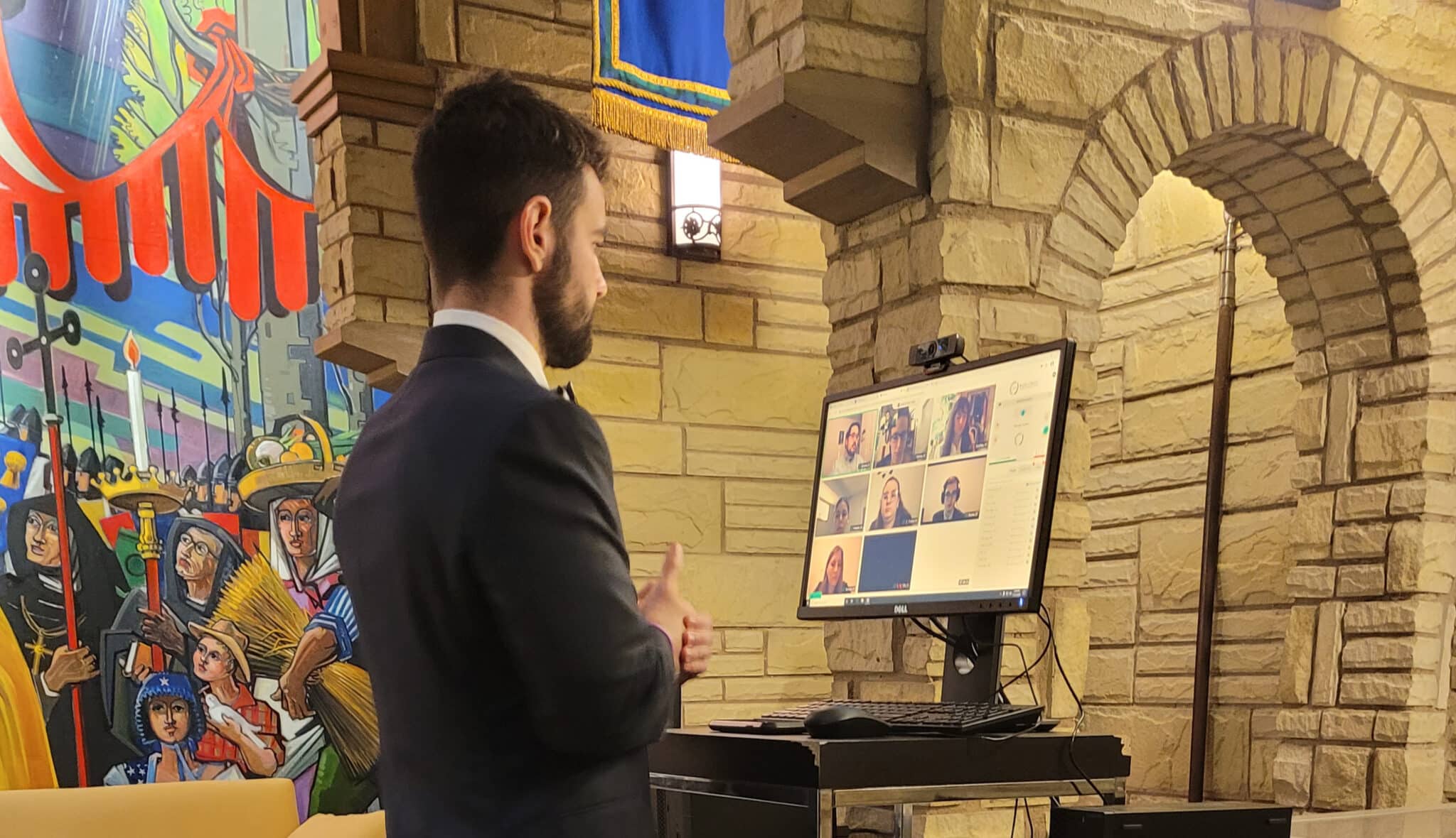 A man in a suit presenting in front of a screen displaying a grid of video call participants, in Howard Payne University, with colorful wall art. | HPU