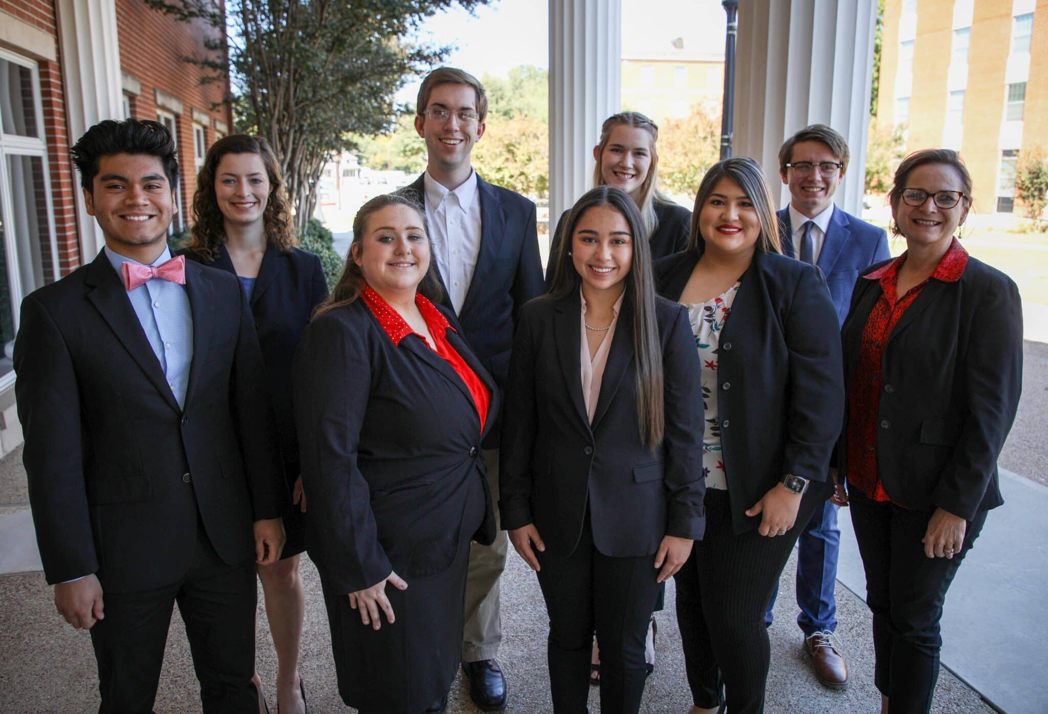 Group of eight individuals posing for a photo on Howard Payne University's entrance, dressed in professional attire. | HPU