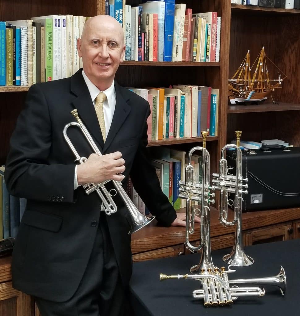 Man holding a trumpet standing next to a table with two additional trumpets on display at Howard Payne University. | HPU