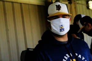 A person wearing a white face mask and a baseball cap with the letters "nd" from Howard Payne University stands in a shaded area. | HPU