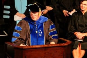 An academic figure in Howard Payne University regalia speaking at a podium during a graduation ceremony. | HPU
