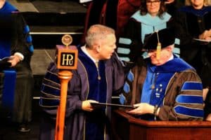 An academic ceremony at Howard Payne University with two individuals in regalia, one handing a document to the other by a podium with the university emblem. | HPU