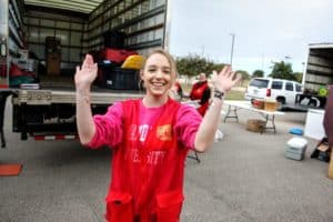 A smiling woman in a red shirt with raised hands gestures a playful pose at an outdoor Howard Payne University event with another person and a truck in the background. | HPU
