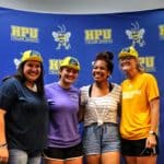 Four individuals smiling in front of a banner with the Howard Payne University logo, three of them wearing yellow construction hats. | HPU