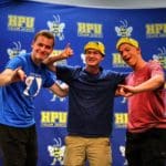 Three individuals posing with thumbs up in front of a backdrop with Howard Payne University logo emblems. | HPU