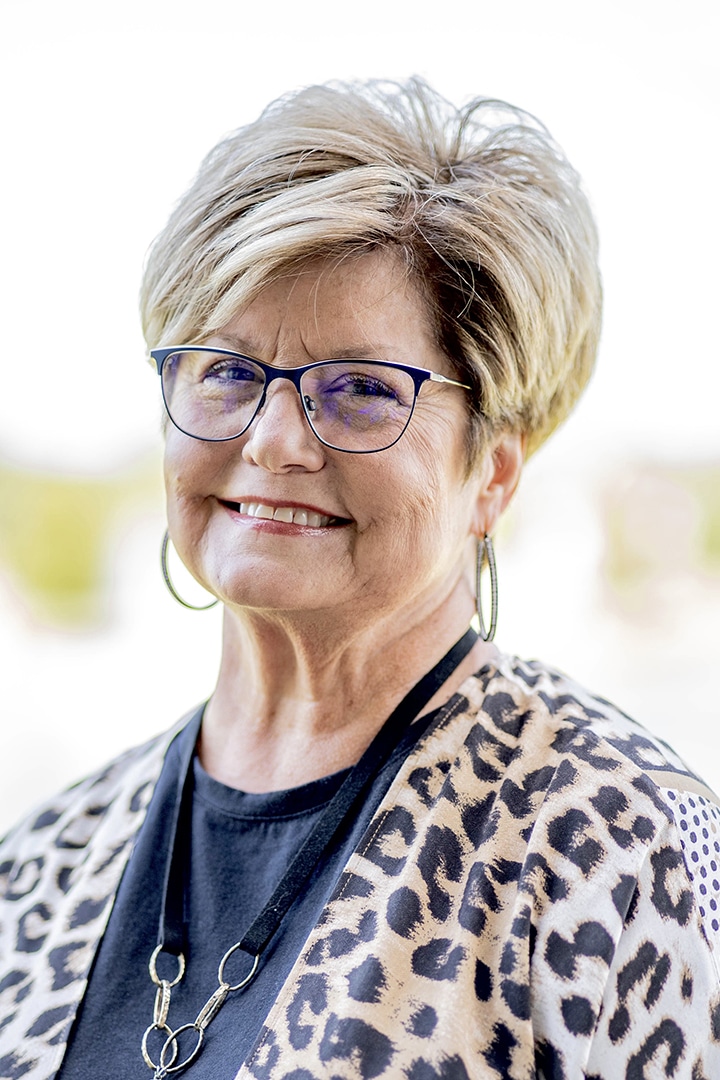 A smiling woman with short hair wearing glasses, hoop earrings, a necklace, and a Howard Payne University leopard print top. | HPU