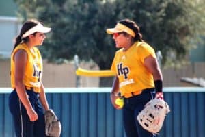 Two Howard Payne University softball players in yellow jerseys conversing on the field. | HPU