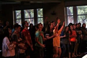 Group of individuals with raised hands attending an indoor event at Howard Payne University, possibly a concert or worship service, with dim lighting. | HPU