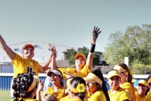 A Howard Payne University softball team in yellow uniforms celebrates a victory on the field, with one player raising her hand in triumph. | HPU