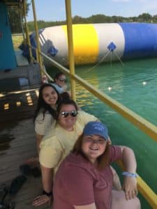 Three individuals smiling on a dock with a large blue and yellow inflatable Howard Payne University structure in the water behind them. | HPU