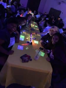 Group of Howard Payne University people smiling at a table with electronic devices in a dimly lit room. | HPU