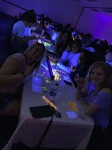 A group of Howard Payne University students enjoying a glow-in-the-dark event with smiles around a table illuminated by neon lights. | HPU