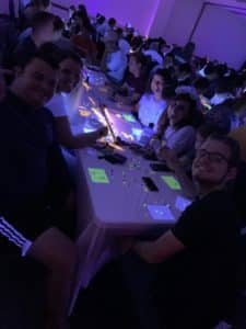 Group of Howard Payne University students sitting at a table with glowing decorations in a dimly lit room, smiling at the camera. | HPU
