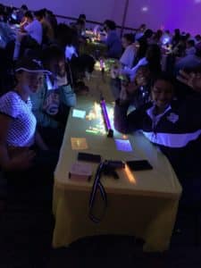 Group of Howard Payne University students sitting at tables with glowing decorations in a dimly lit room, smiling and waving at the camera. | HPU
