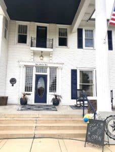 Two-story white house with a blue front door, a small balcony above, and a sign indicating Howard Payne University is open. | HPU