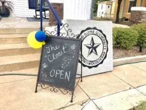 A chalkboard sign reading "the closet open" with balloons attached, standing next to a stone sign for Howard Payne University - alumni woods. | HPU