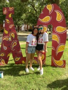 Two individuals posing with large Greek letter cutouts outdoors at Howard Payne University. | HPU