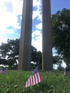 Small american flags planted in grass at Howard Payne University with large columns in the background and a blue sky with clouds above. | HPU