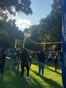 Outdoor event at Howard Payne University with people gathered on a grassy area, some playing volleyball, with trees and sunlight in the background. | HPU