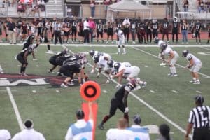 Two Howard Payne University American football teams lined up at the line of scrimmage during a game, with referees and sideline players in the background. | HPU