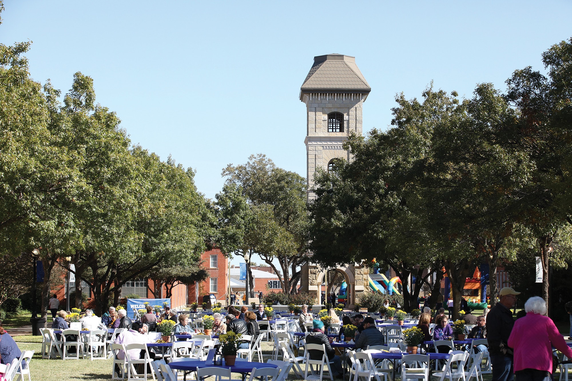 Outdoor event with guests seated at tables in a park with Howard Payne University's tower in the background. | HPU