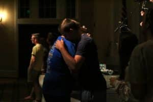 Two individuals embracing in a dimly lit room with others around at Howard Payne University. | HPU