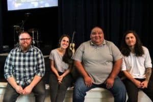 Four people smiling and posing together on a stage at Howard Payne University with musical instruments in the background. | HPU