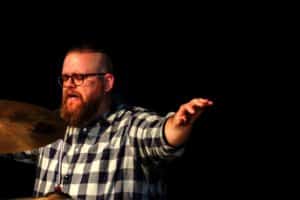 A Howard Payne University man with glasses wearing a plaid shirt playing the drums on stage. | HPU