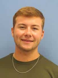 A young man with short brown hair smiling at the camera, wearing a Howard Payne University green t-shirt and a necklace, against a blue background. | HPU