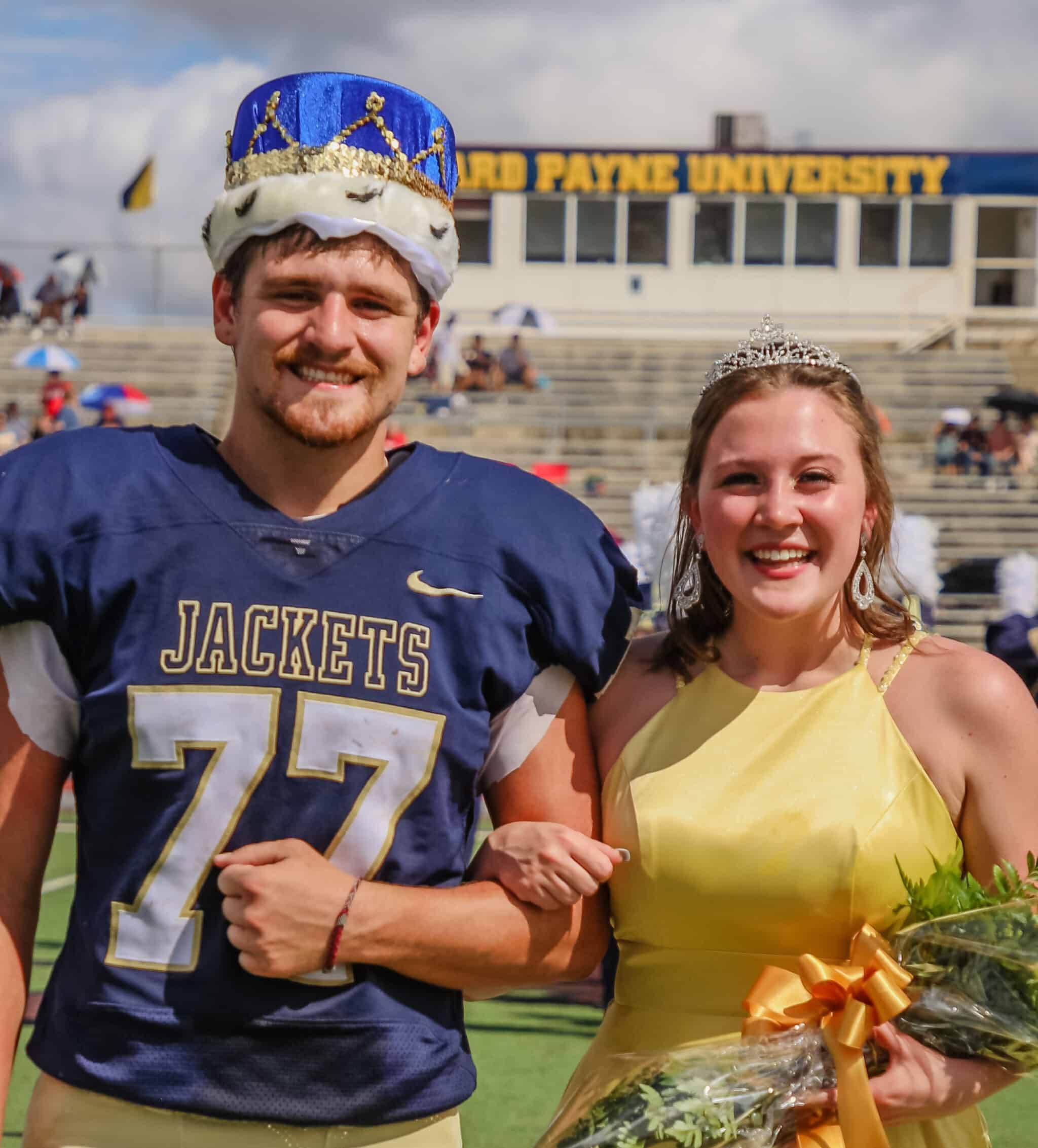 A football player from Howard Payne University and a young woman wearing a yellow dress and tiara, both with crowns, smiling on a sunny day at a stadium. | HPU