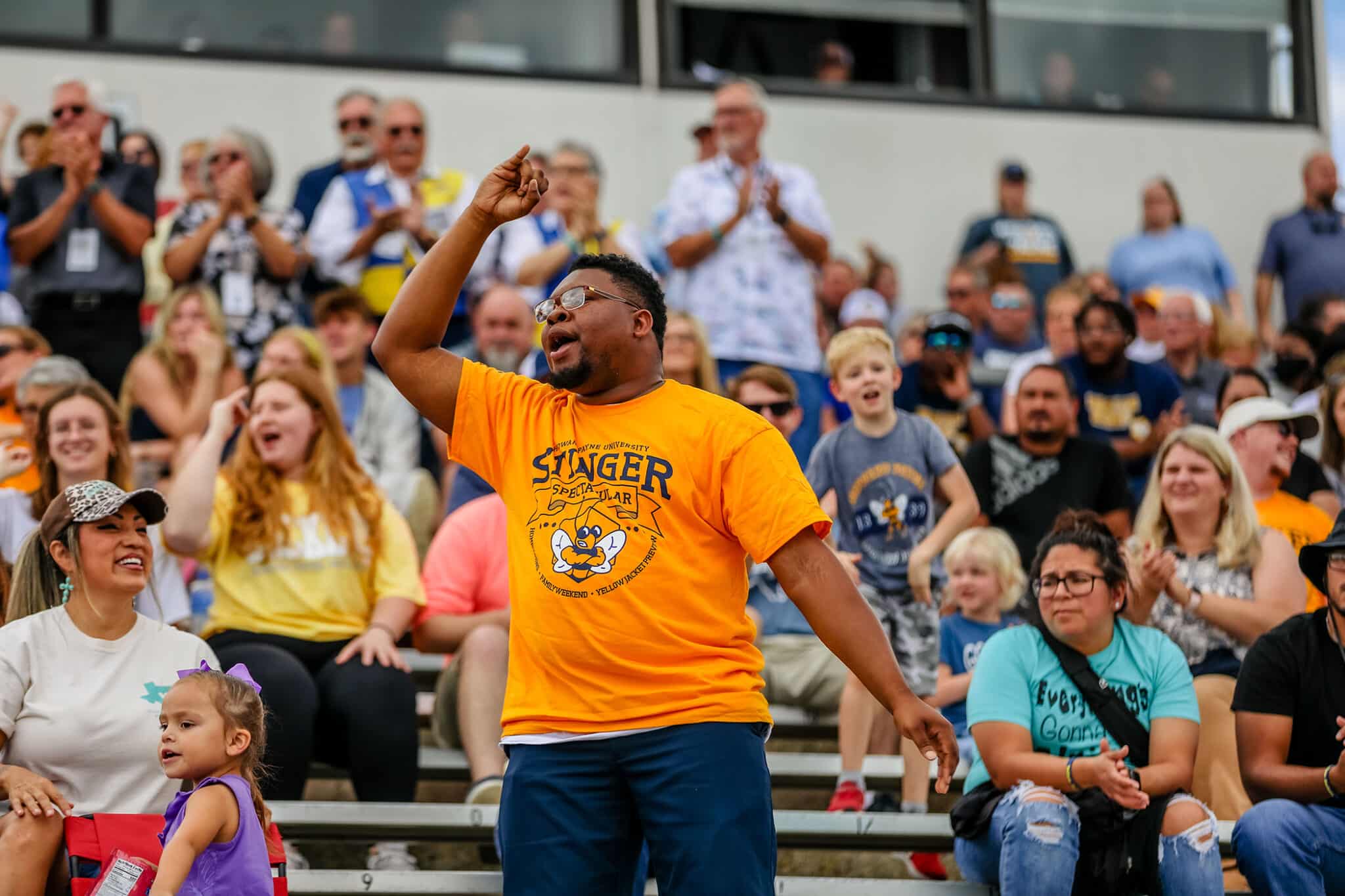 A man in a yellow Howard Payne University t-shirt cheering at an event while surrounded by an audience in the stands. | HPU