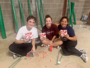 Three Howard Payne University students sitting on the floor holding beverage cups and smiling, with various painting supplies around them. | HPU