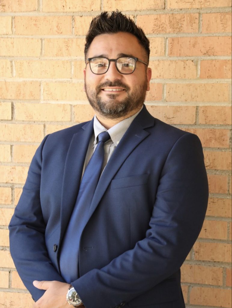 A confident man in Howard Payne University business attire is standing against a brick wall. | HPU