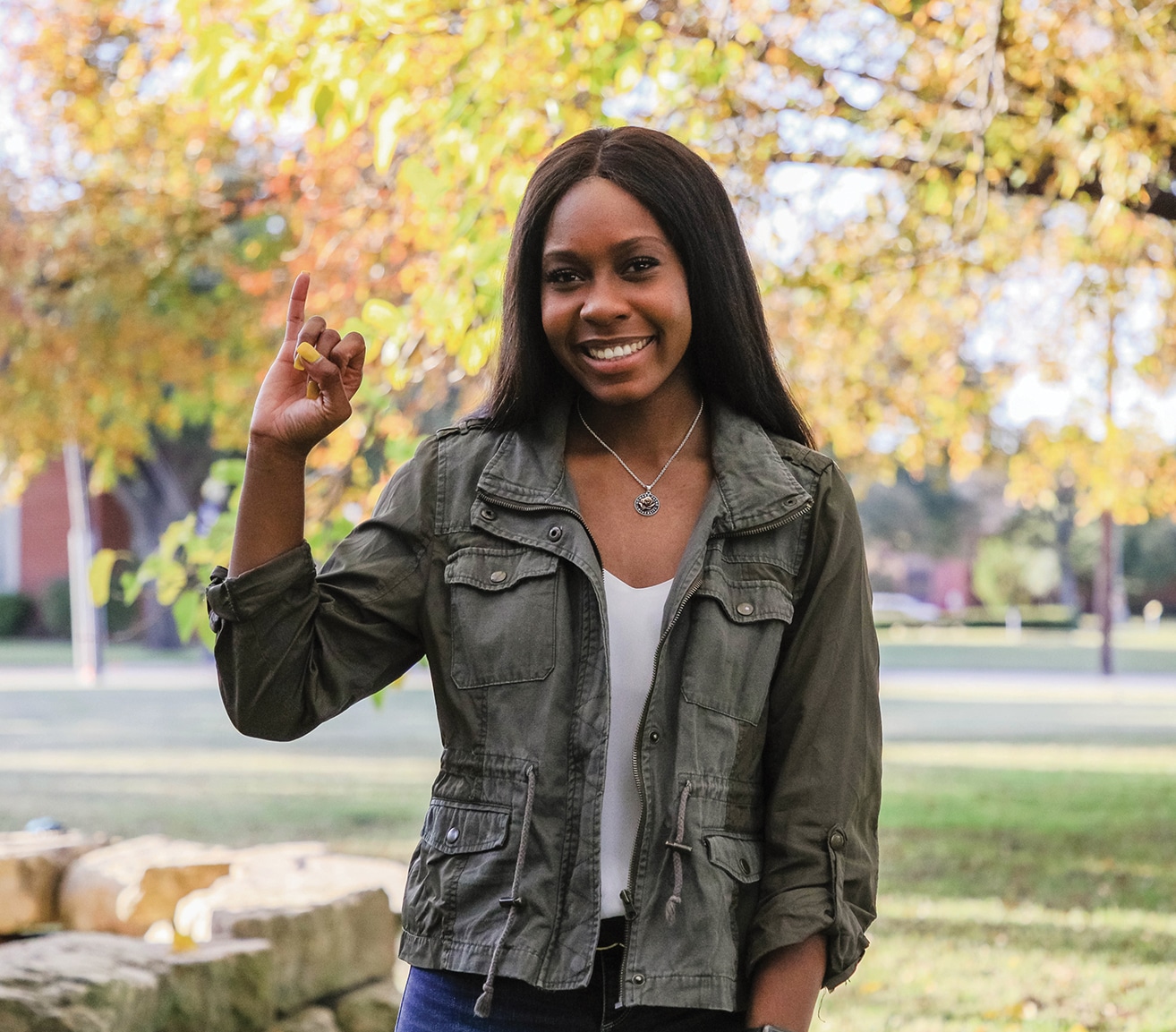 A smiling woman giving a peace sign outdoors with Howard Payne University's trees in the background. | HPU