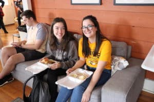 Two individuals smiling at the camera while sitting on a couch, holding food on plates at Howard Payne University. | HPU