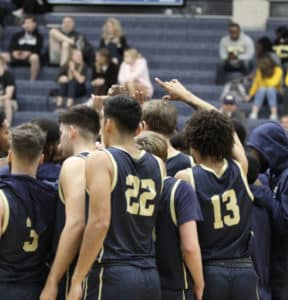 A Howard Payne University basketball team in a huddle with one player raising his hand, on the court with spectators in the background. | HPU