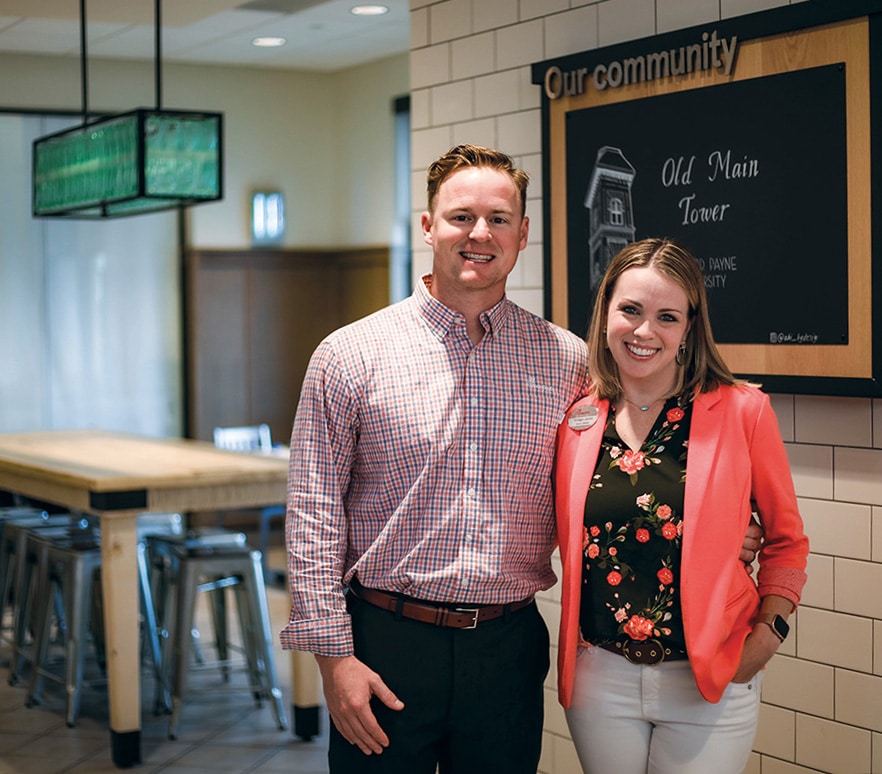 Two individuals smiling in a casual office setting at Howard Payne University with a community bulletin board in the background. | HPU