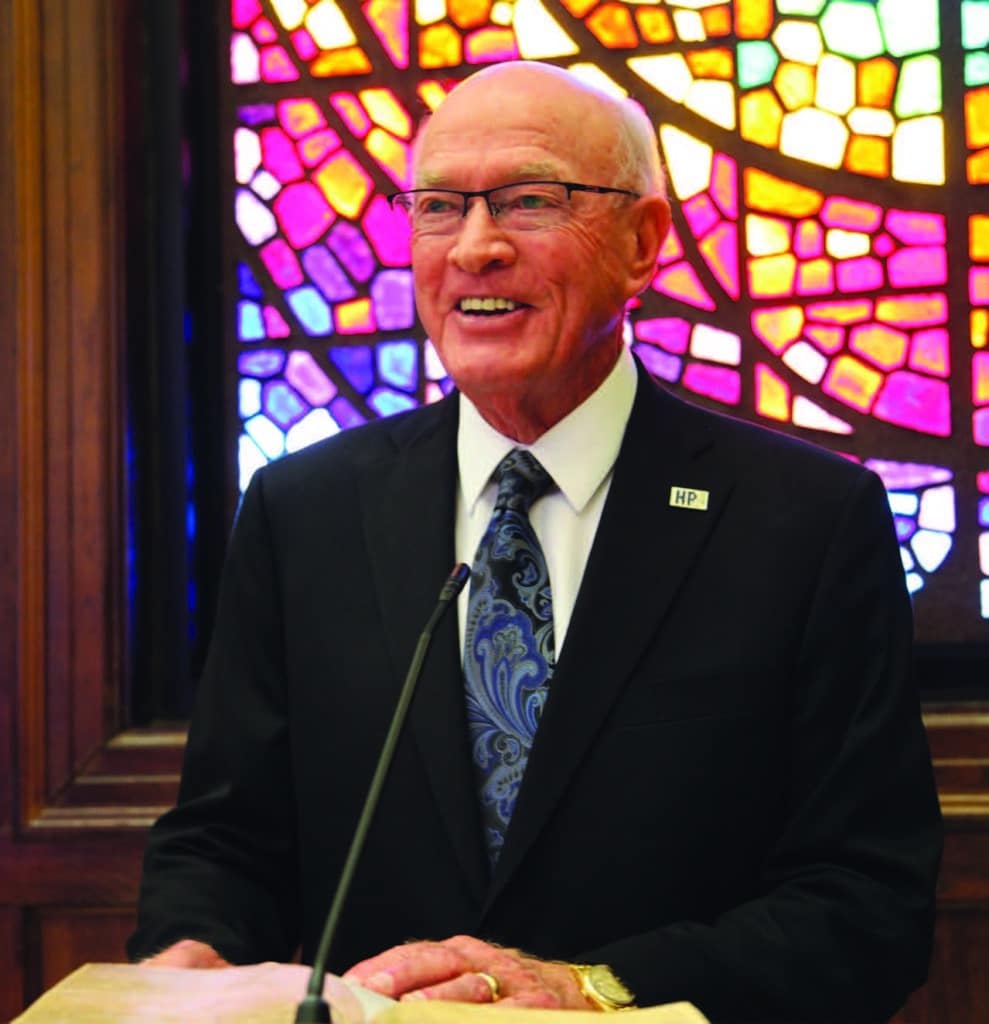 A smiling man in a suit and tie speaking at a podium with a colorful stained-glass window in the background at Howard Payne University. | HPU