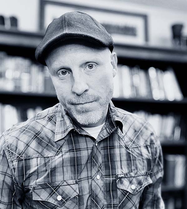 A man in a cap and plaid shirt posing for a portrait with Howard Payne University bookshelves in the background. | HPU