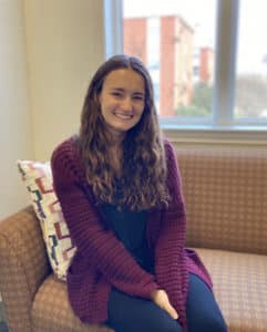 A smiling woman sitting on a couch by the window, wearing a Howard Payne University purple cardigan. | HPU