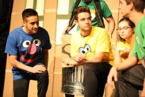 Four individuals in colorful shirts with Howard Payne University logos and cartoon faces printed on them engaged in a group activity or performance, with one man playing a steel drum. | HPU