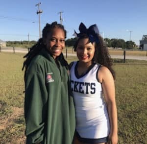 Two individuals posing together, one wearing a cheerleading outfit from Howard Payne University and the other in a green jacket, with an outdoor setting in the background. | HPU