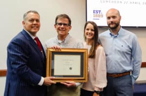 Four individuals posing for a photo, one holding a framed certificate from Howard Payne University, in a room with a sign indicating an event held on April 29. | HPU