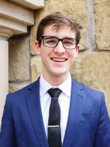 Young man in a blue suit and tie smiling against a stone wall background at Howard Payne University. | HPU